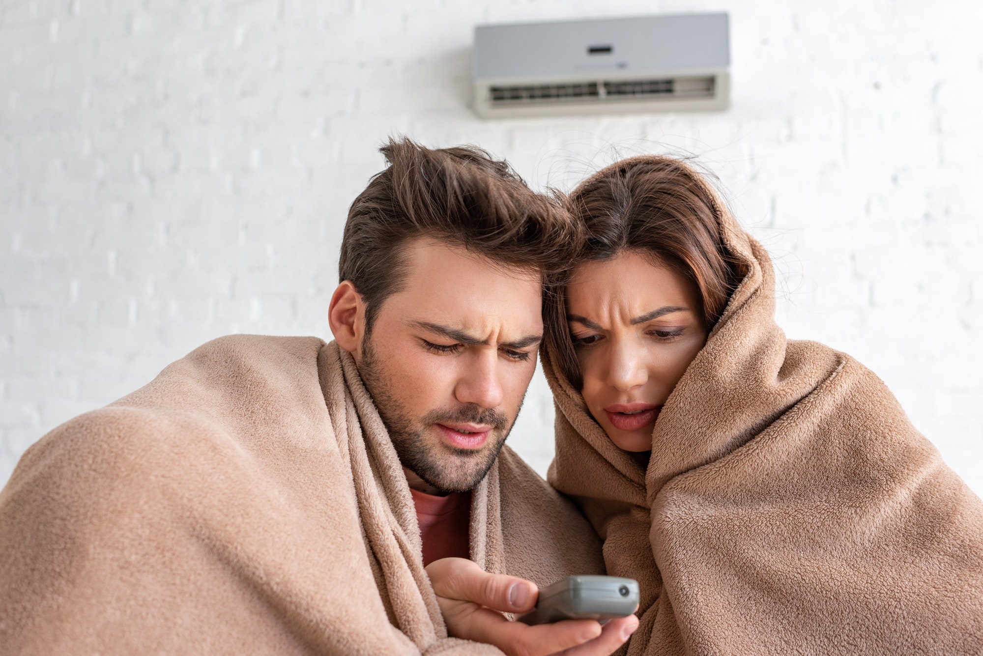frozen man and woman warming under blankets while sitting under air conditioner with remote control